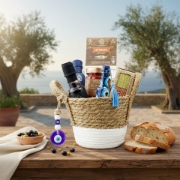 Gourmet Greek gift basket displayed on a wooden table with olives, bread, and olive trees in the background overlooking the sea.