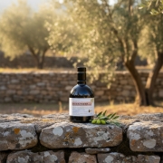 Organic extra virgin olive oil bottle resting on an ancient Greek stone wall with a green olive branch and sun-drenched Athinolia olive trees in the background.