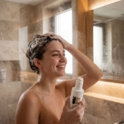 A smiling woman using the Mastic Spa 2 in 1 Shampoo and Conditioner in a modern bathroom shower, showing the rich lather in her hair while holding the product bottle.